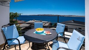 A round table with a bowl of fruit and watermelon slices, surrounded by blue chairs with matching cushions, overlooking a clear blue sea and distant hills.