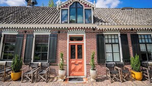 A brick building with a red door, black shutters, and a tiled roof. There are outdoor seating areas with chairs and potted plants. - Hotel Suites Wilma J (Vinkeveen)