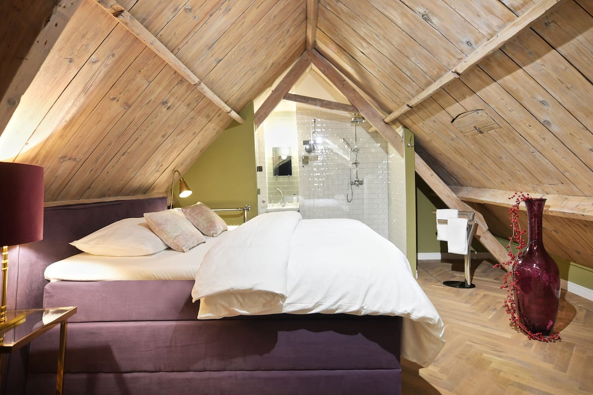 Une chambre avec un plafond en pente, un lit, une table de chevet, une lampe, un vase et un espace douche.