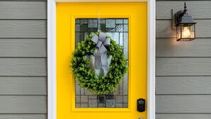 Una puerta amarilla brillante con un panel de vidrio, un adorno de guirnalda, un timbre y una lámpara fijada a la pared.