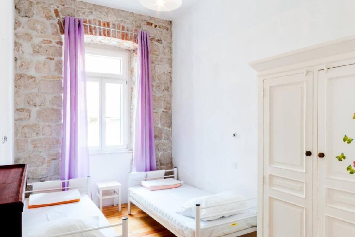 A bedroom with a stone wall, a window with purple curtains, a white wardrobe, and a bed with a white mattress and pillows.