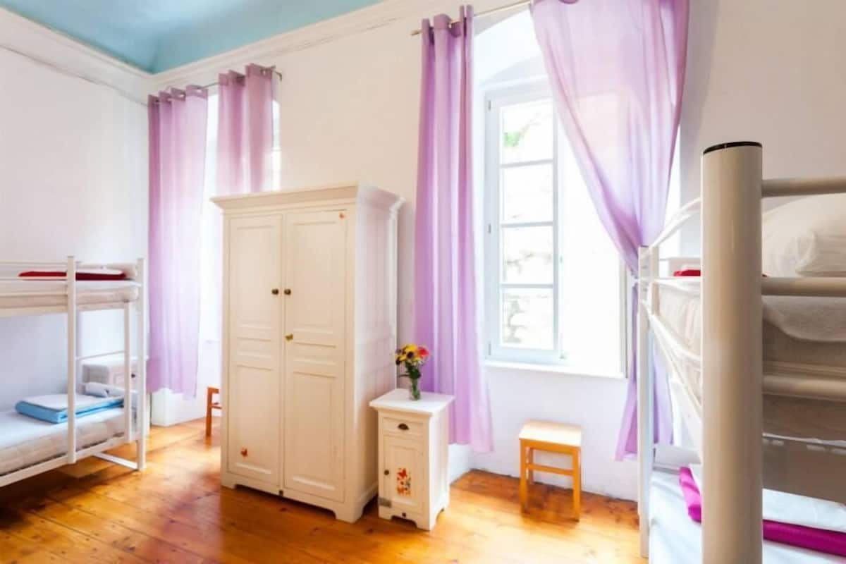 A bedroom with bunk beds, a wardrobe, a small table with a vase, and a window with purple curtains.