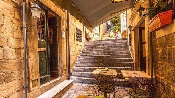 A narrow alleyway with stone walls, a set of stone stairs leading to a higher level, outdoor tables and chairs, and a door with a number.