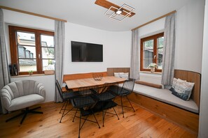 A room with a wooden dining table, black wire chairs, a grey sofa, and a TV mounted on the wall.