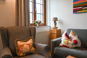 Ein Wohnzimmer mit zwei Sesseln, einer Heizung, einem Fenster mit Vorhängen, einer Vase mit Blumen und einem farbenfrohen abstrakten Gemälde an der Wand.