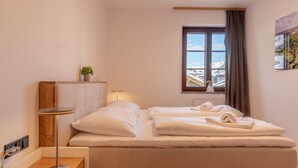 Ein Hotelzimmer mit Bett, Nachttisch, Lampe und einem Fenster mit Blick auf Gebäude und Berge.