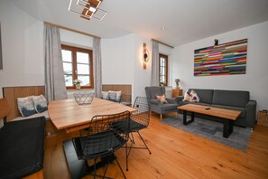 A modern living room with a wooden dining table, black wire chairs, a grey sofa, and a colorful abstract painting on the wall.