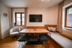 Ein Essbereich mit einem Holztisch, vier Stühlen, einer Couch, einem an der Wand montierten Fernseher und einem fenster mit Vorhängen.