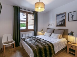 A bedroom with a bed, a chair, a bedside table, a lamp, a window with curtains, and a wall-mounted picture.