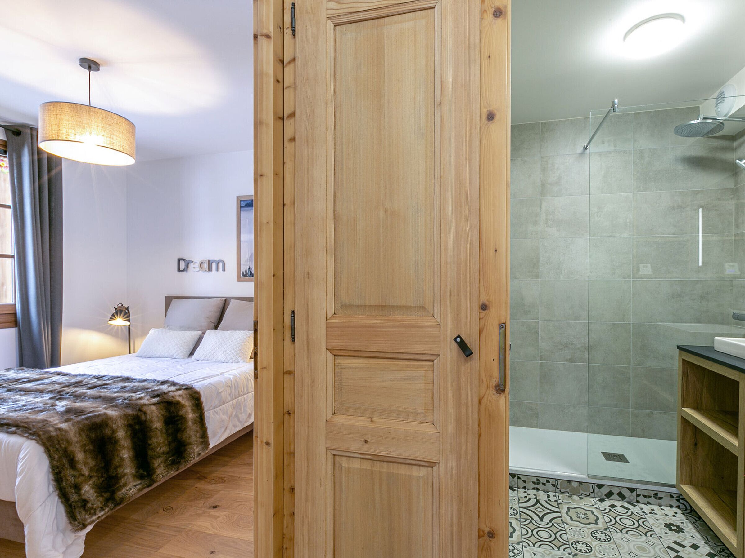 A bedroom with a bed, a bedside lamp, and a wooden door leading to a bathroom with a shower and tiled walls.