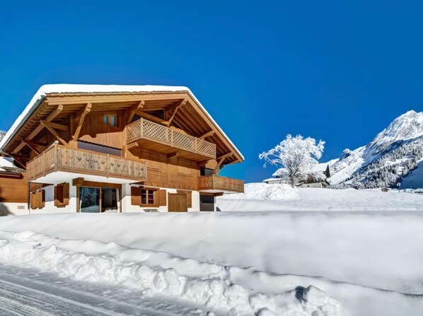 A chalet with a wooden balcony and a snow-covered roof, set against a mountainous backdrop.