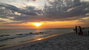 En strand med fodaftryk i sandet, en solnedgang i baggrunden og mennesker på kysten.