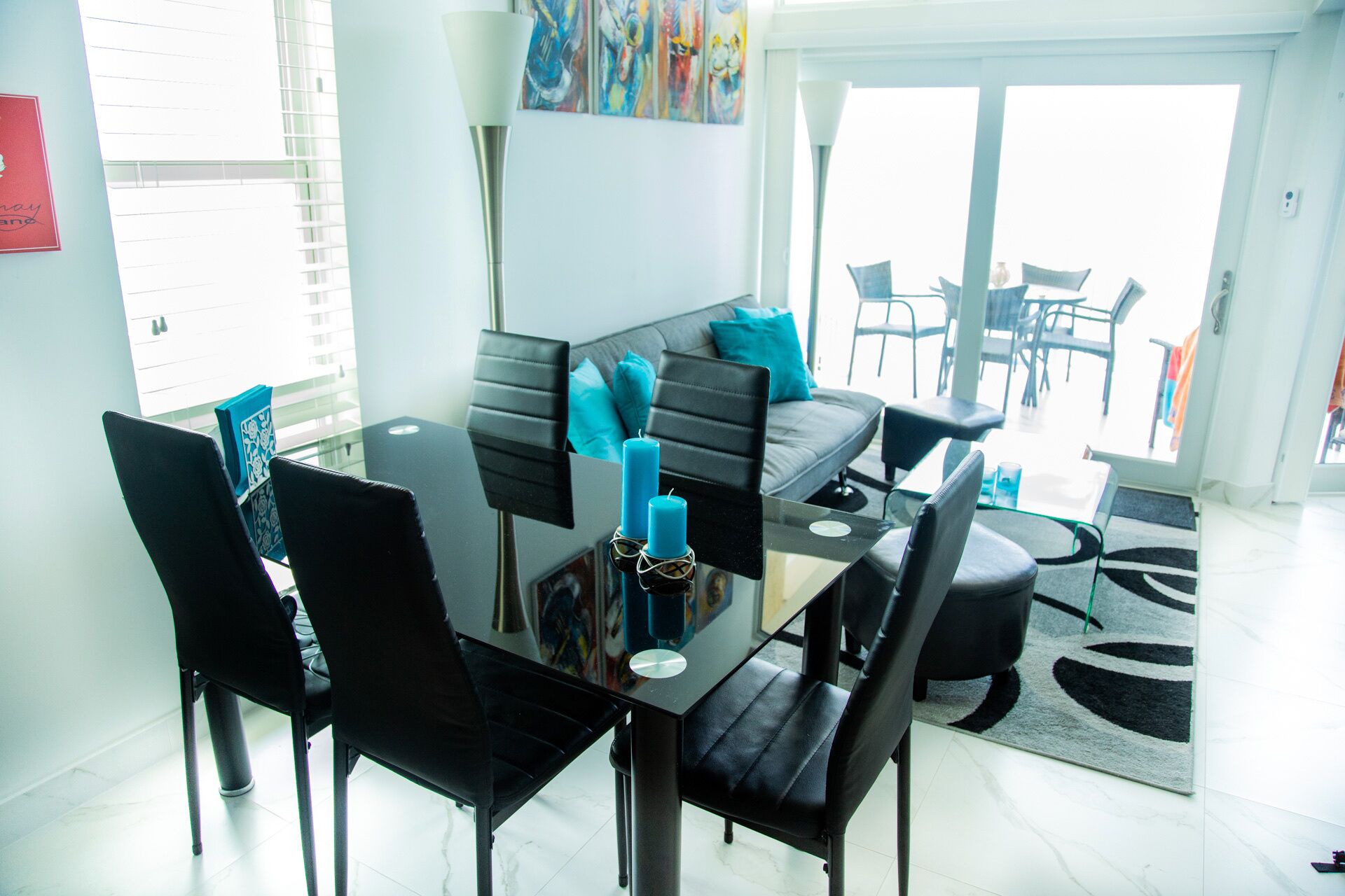 A modern dining area with a glass table, black chairs, and a grey sofa. There are two blue candles on the table.