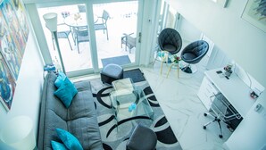 Una sala moderna con un sofá gris, una mesa de centro de vidrio y una silla de oficina negra. Se ve una ventana grande con vistas a muebles de exterior.