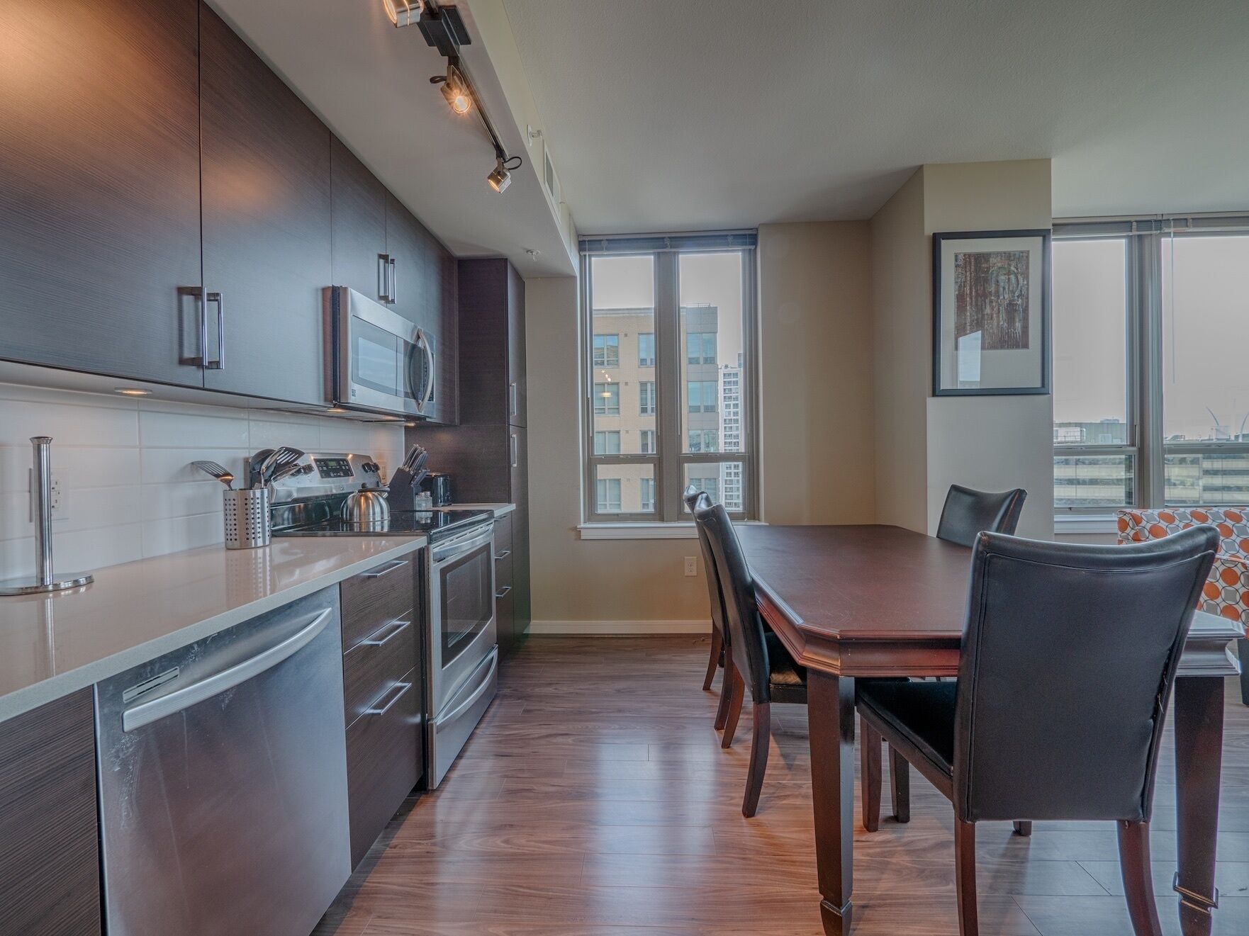 A modern kitchen with stainless steel appliances, a dining area with a wooden table and chairs, and a large window offering a city view.
