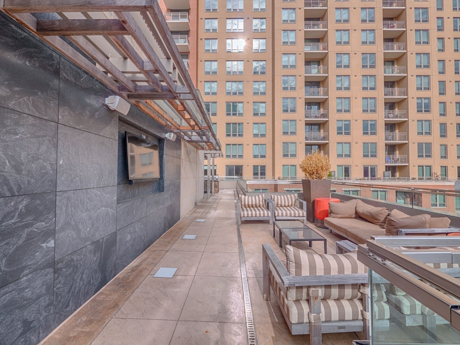 A rooftop terrace with outdoor seating, a TV mounted on the wall, and a view of a high-rise building in the background.
