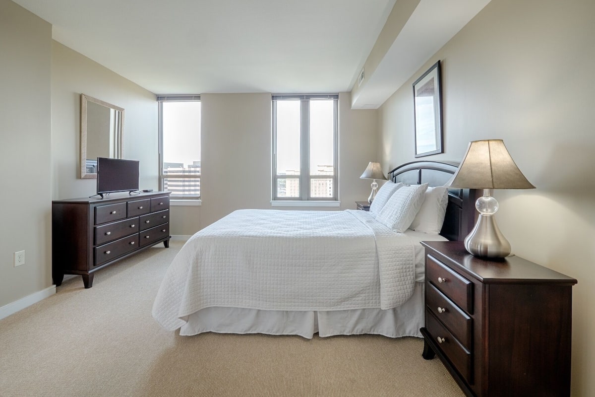 A bedroom with a large bed, a dresser, a TV, and two windows.