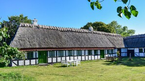 Une maison traditionnelle à toit de chaume, avec une façade à pans de bois peinte en blanc et en noir, une porte verte et un petit espace extérieur aménagé pour s’asseoir.