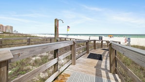 Ein hölzerner Steg führt zu einem Strand mit einer Flagge, einem sandigen Bereich und Gebäuden im Hintergrund.