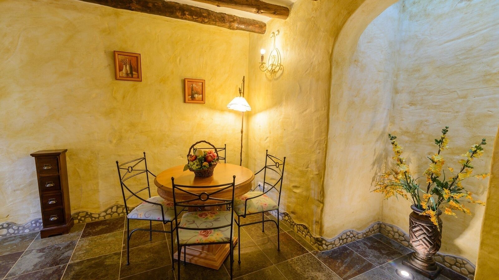 A dining area with a round table, floral chairs, a vase with flowers, and a wall-mounted lamp.