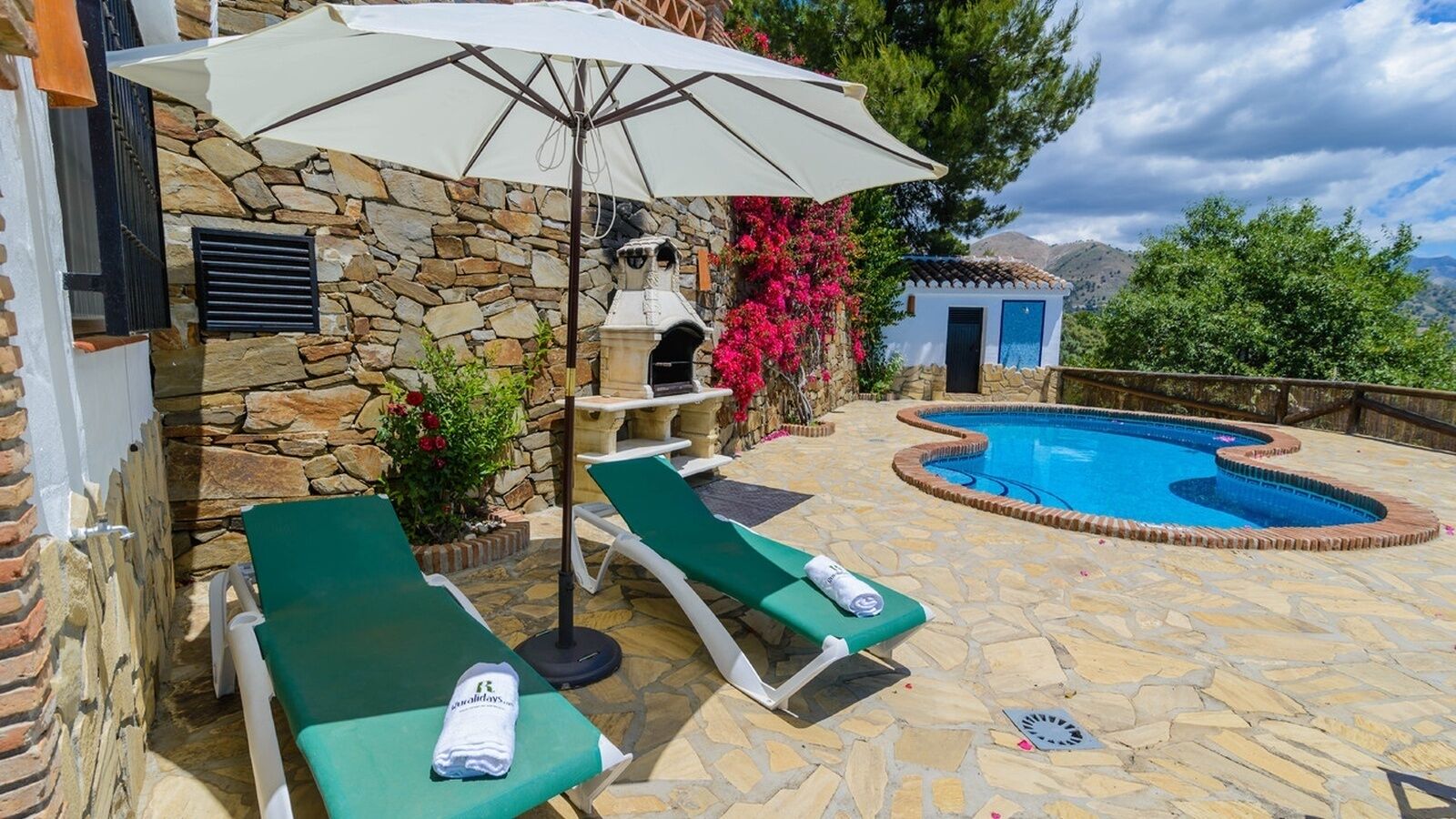 A stone patio with two green lounge chairs and a white umbrella, a small pool with a stone border, and a stone wall with a chimney.