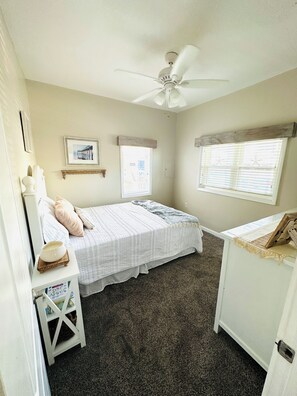 Un dormitorio con una cama, una mesita de noche, un aparador, un ventilador de techo, dos ventanas con persianas y un cuadro enmarcado en la pared.