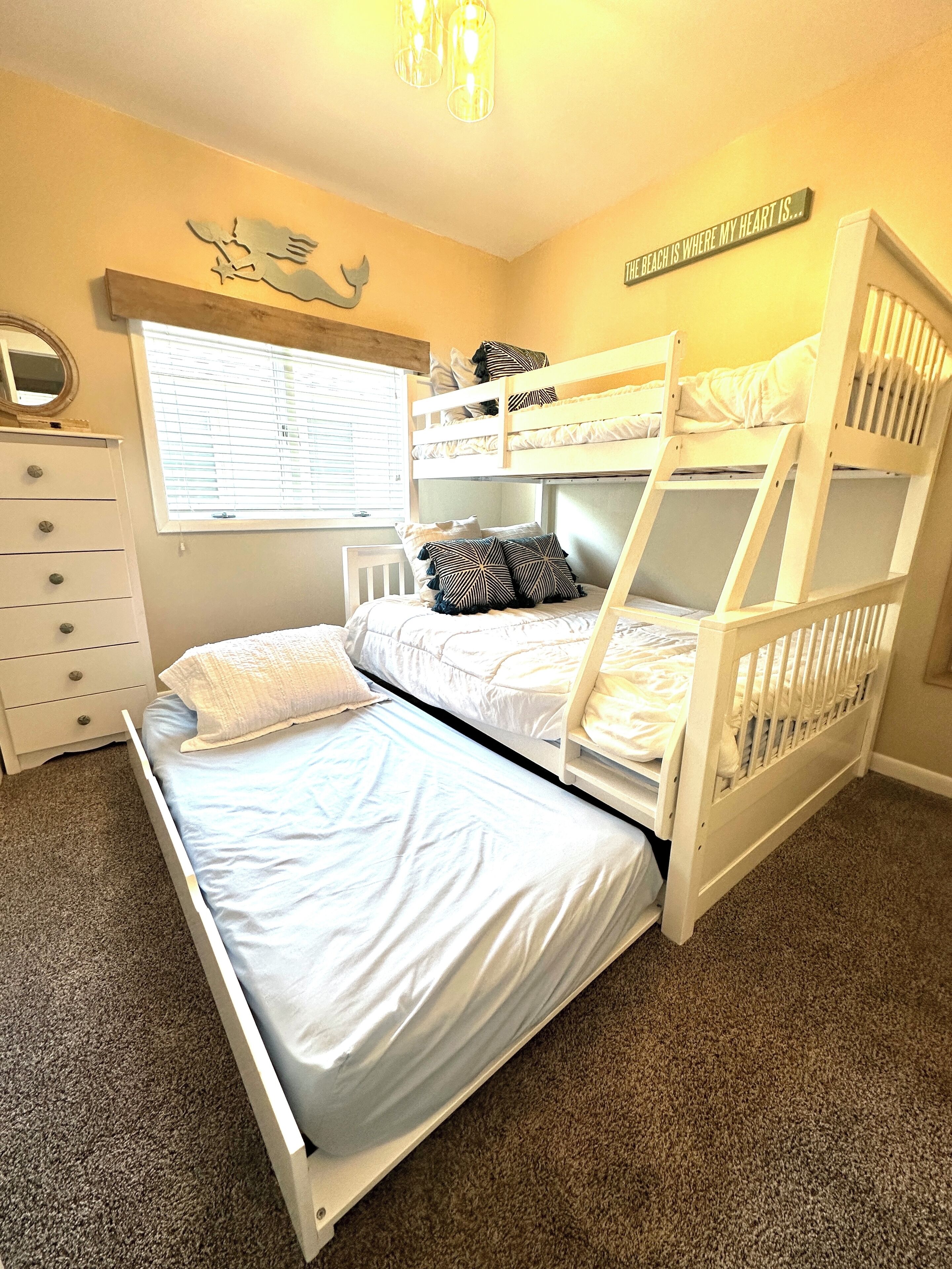 A bunk bed room with a dresser, a window with blinds, and a wall decoration.