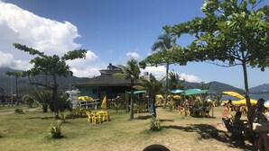 A beachside restaurant with outdoor seating, palm trees, and a mountainous backdrop.