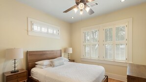 A bedroom with a wooden bed, two bedside tables with lamps, a ceiling fan, and windows with shutters. - Hyde Park Historic District - Close To Everything Austin Has To Offer! (Austin)