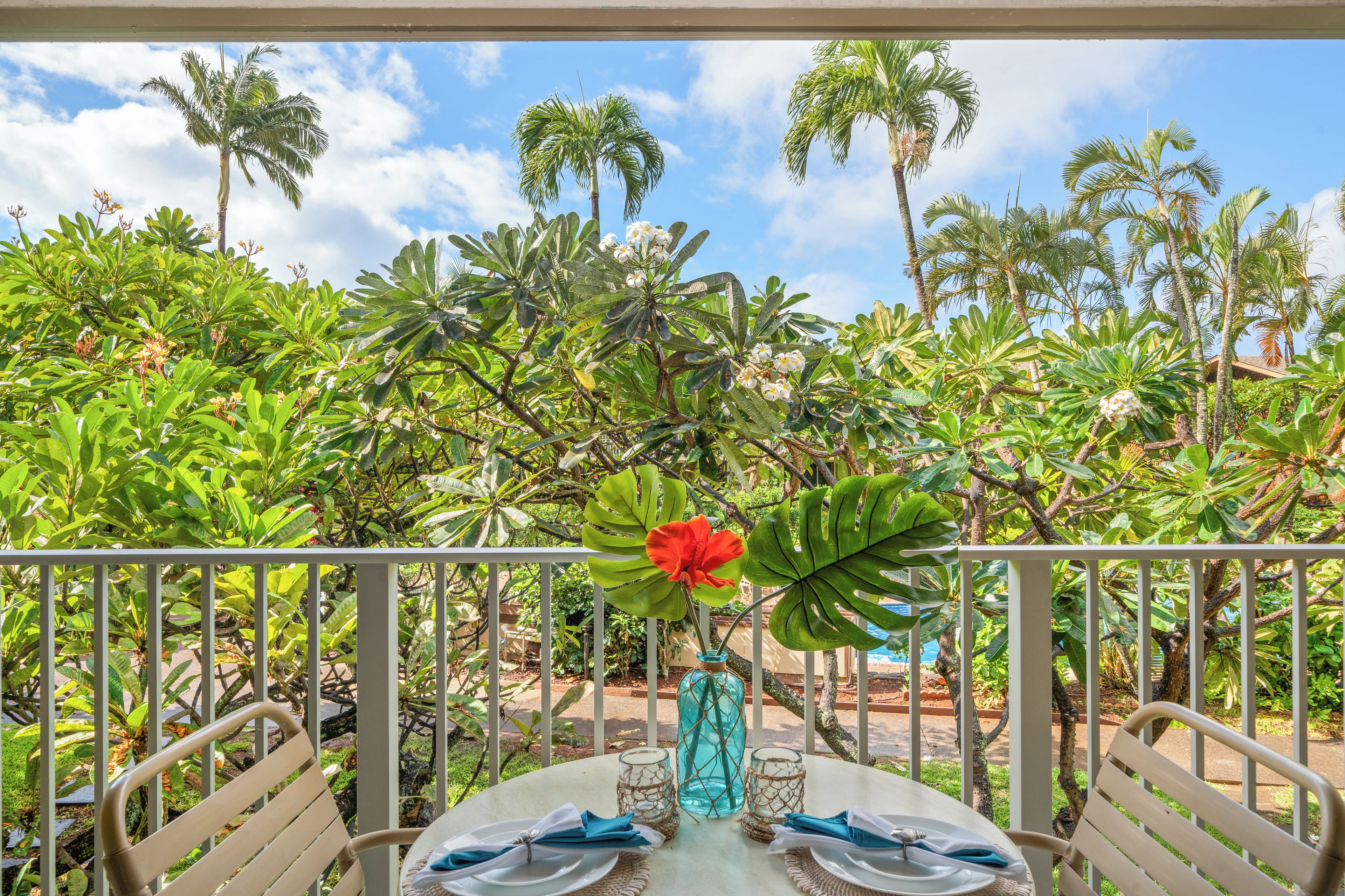 Une table dressée pour quatre personnes, agrémentée d’un vase de fleurs, entourée d’une végétation tropicale et de palmiers.