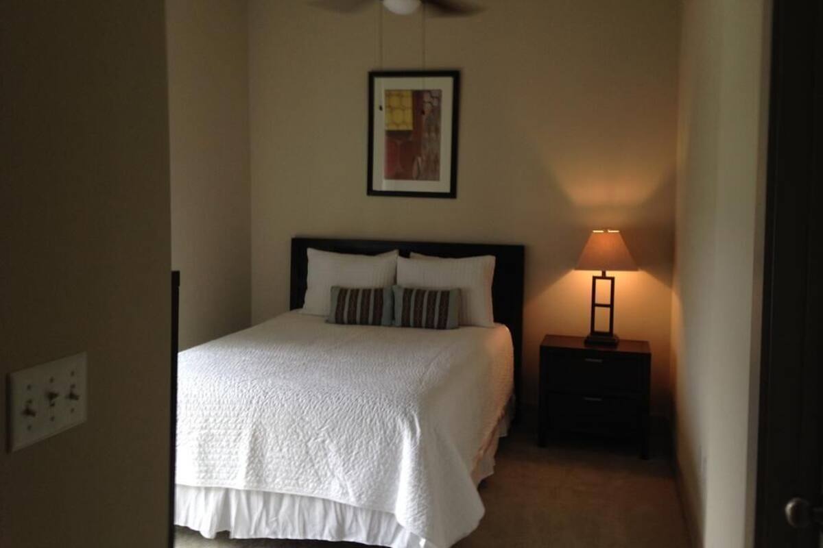 A hotel room with a bed, a ceiling fan, a nightstand with a lamp, and a framed picture on the wall.