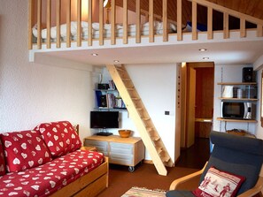 A cozy room with a loft bed, a red patterned sofa, a wooden desk, and a chair with a cushion. - Studio with mezzanine for 2 close to shops and slopes (La Plagne Montchavin Les Coches)