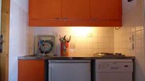 Une cuisine compacte avec des meubles orange, un lave-vaisselle blanc et un petit plan de travail sur lequel se trouve un pot rempli d’ustensiles.