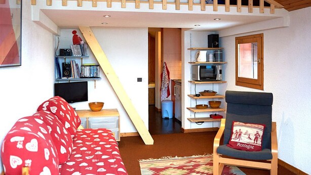 Un salon chaleureux avec un canapé rouge, une chaise en bois et un escalier menant à une chambre à coucher située en mezzanine.