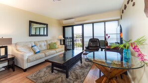 A living room with a sofa, coffee table, and a glass-top table. There is a ceiling fan, a TV, and a window with blinds. - Kihei Akahi Resort, ocean view, steps from the beach (Kihei)
