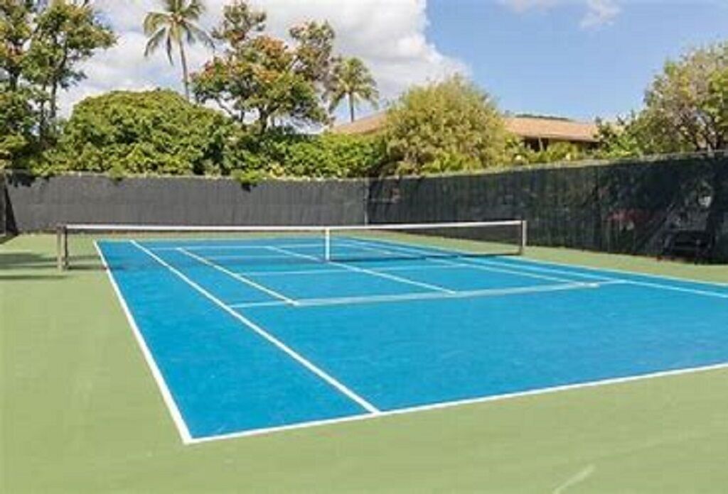 An outdoor tennis court with a net, surrounded by a green fence and trees.
