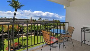 A balcony with a view of a building, a palm tree, and a grassy area. - Kihei Akahi Resort, ocean view, steps from the beach (Kihei)