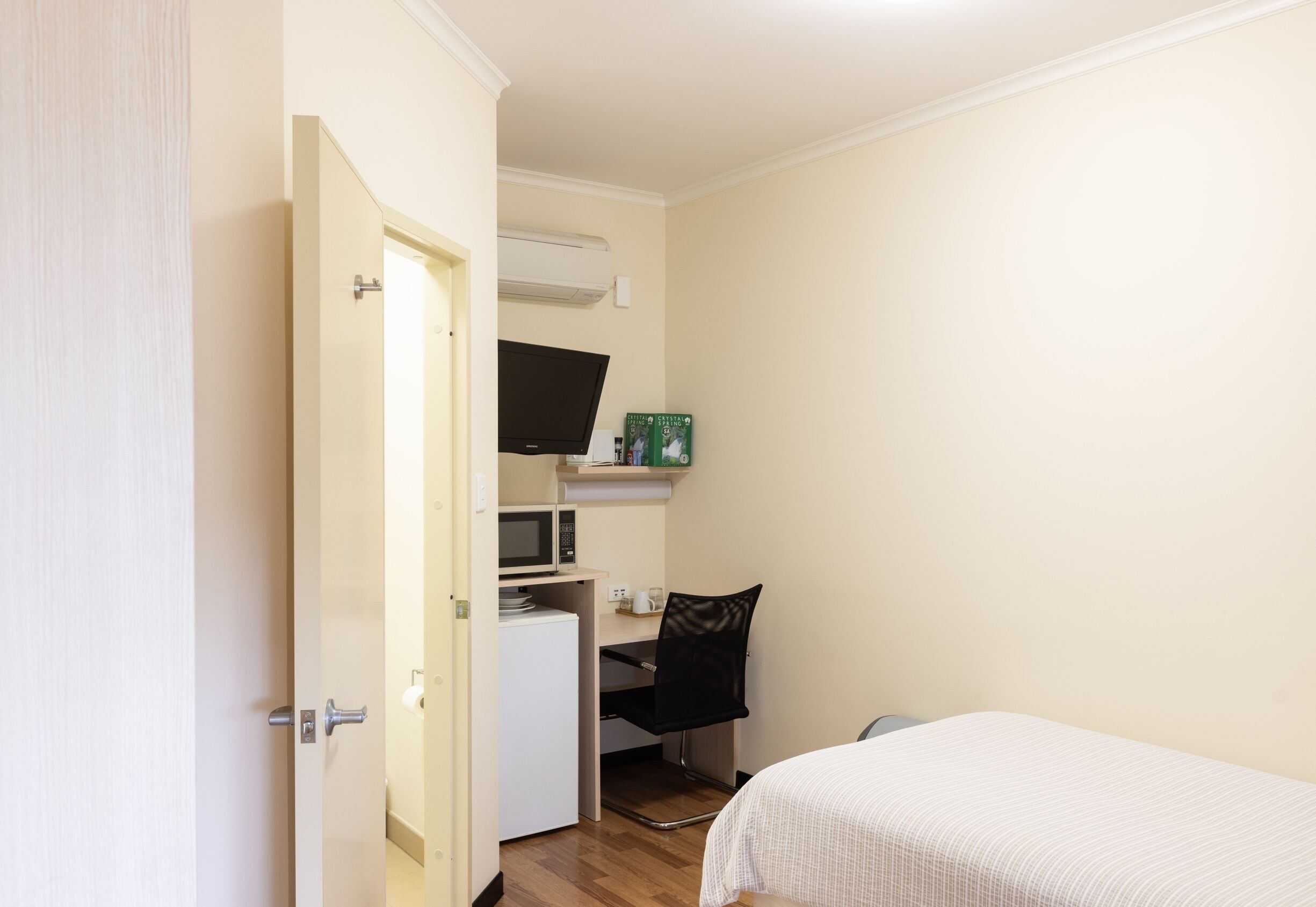 A compact bedroom with a bed, a desk with a chair, a microwave, and a TV on a shelf.