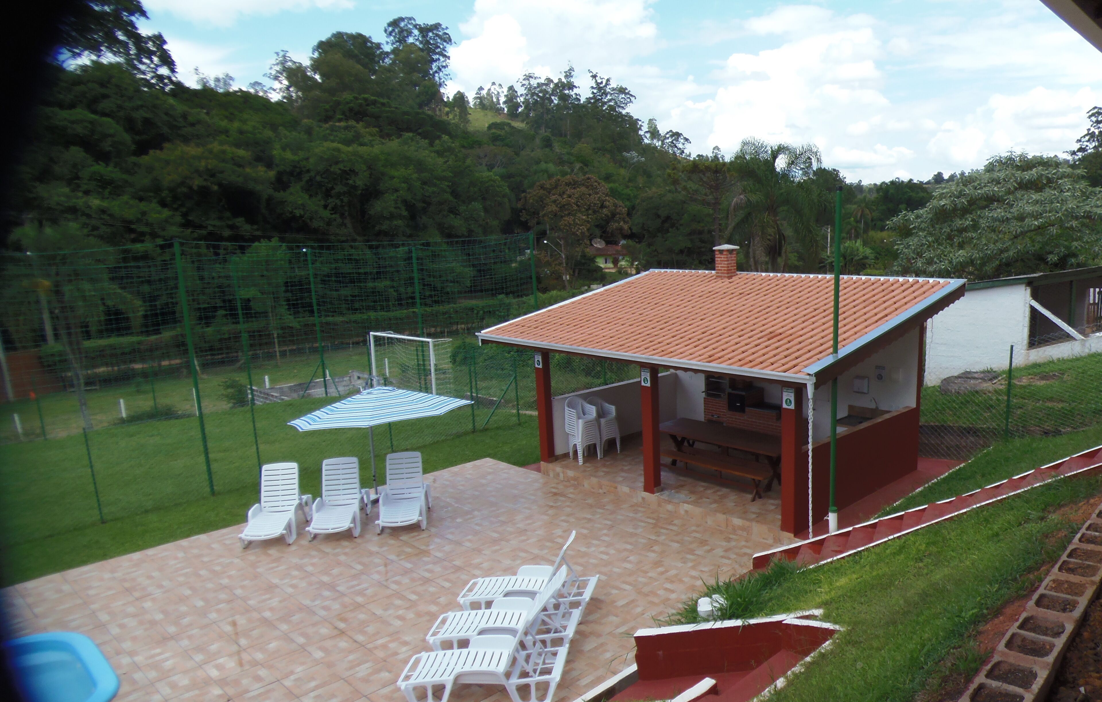 A patio area with white lounge chairs, a small building with a tiled roof, and a fenced garden.