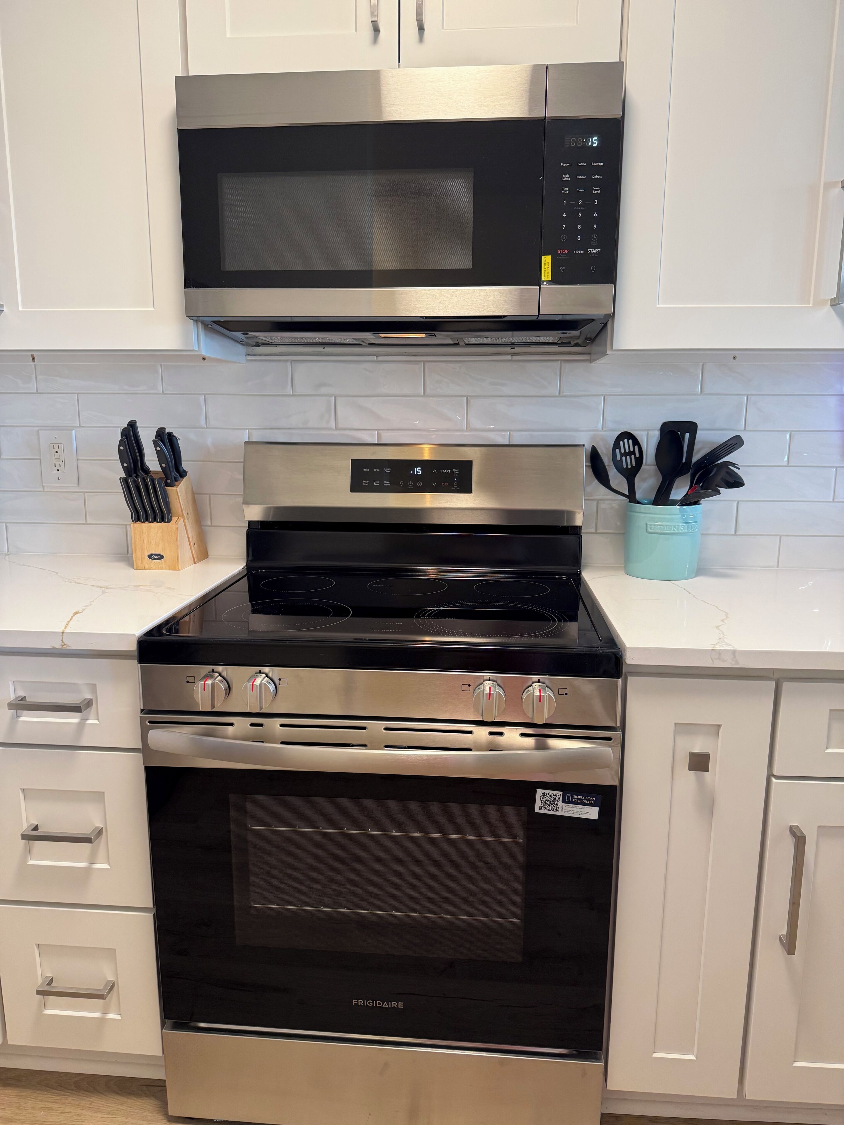 Een keuken met een RVS magnetron, oven en kookplaat, witte keukenkastjes en een houder voor keukengerei op het aanrecht.