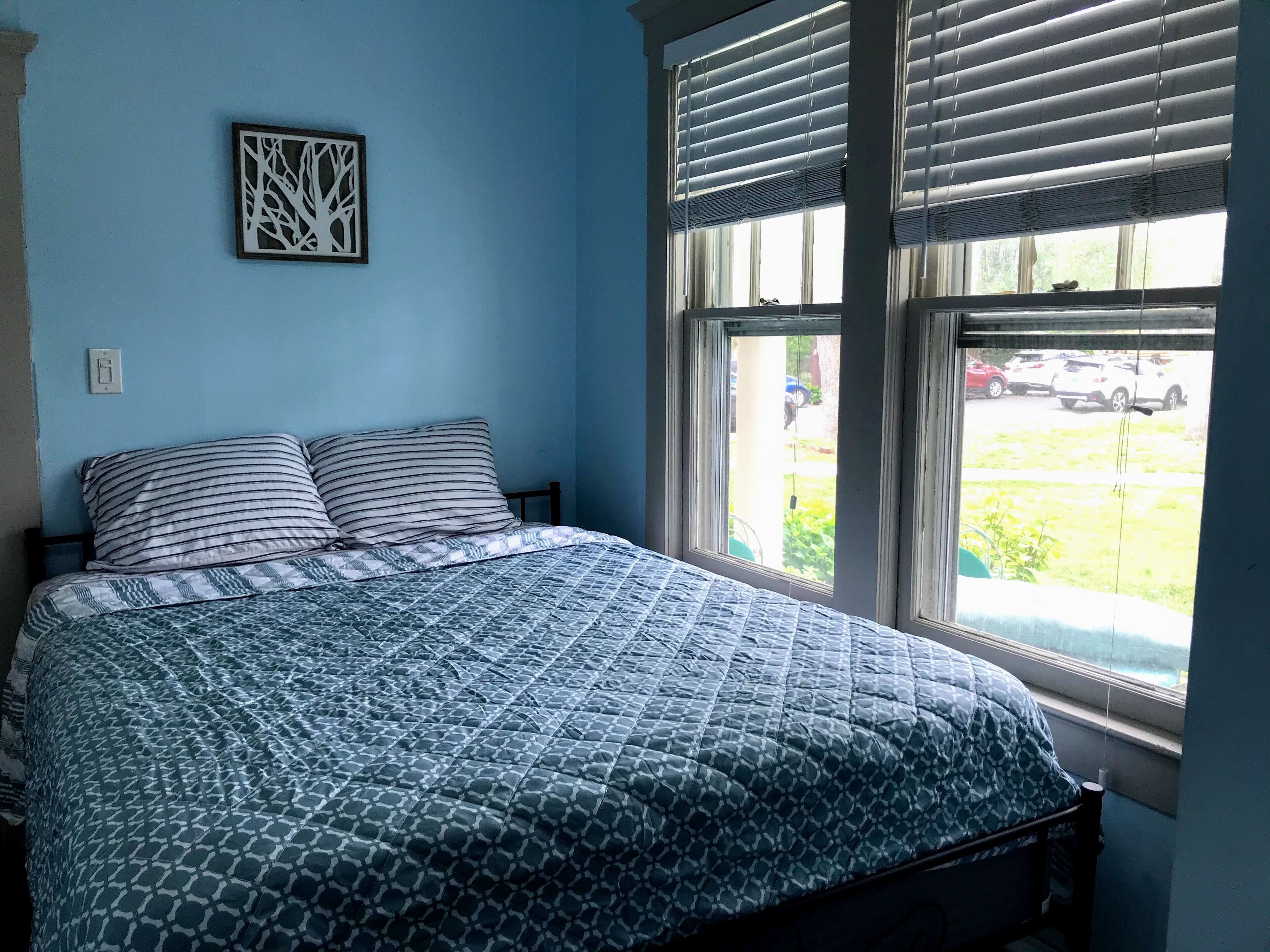 A bedroom with a bed, a window with blinds, and a wall-mounted light switch.