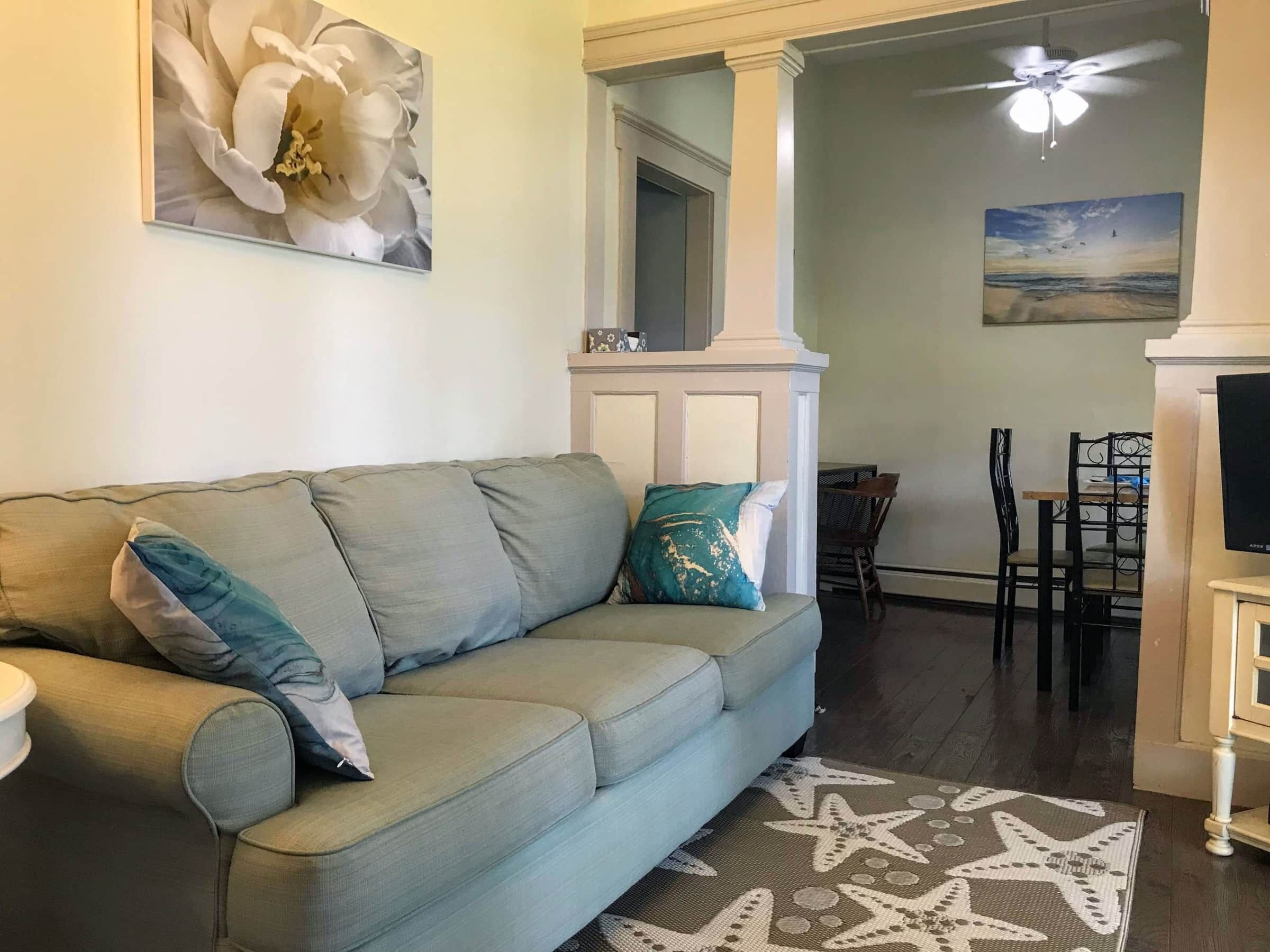 A living room with a light blue sofa, a star-patterned rug, a ceiling fan, and a painting of a beach scene.