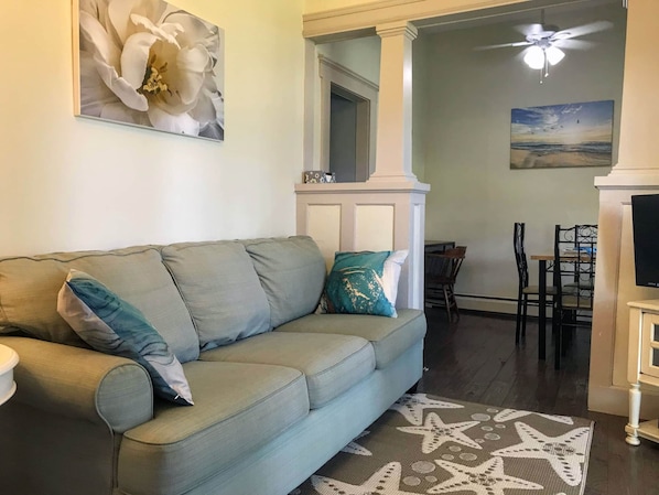 A living room with a light blue sofa, a star-patterned rug, a ceiling fan, and a painting of a beach scene. - Zephyr Retreat Apt 3 - Park View (Neptune Township)