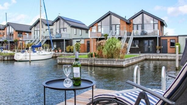 A waterfront view with modern houses, a dock with a sailboat, and a table with a bottle and glass.
