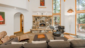 A modern living room with a stone fireplace, leather sofas, and large windows offering a view of the outdoors. - Star Pine_Luxury w/Game Room, Pool Table & HOA Amenities (Hot Tub/Sauna/Gym) (Truckee)