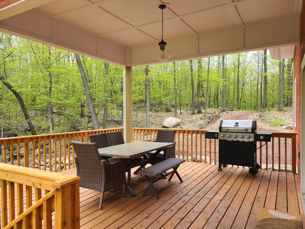 A covered deck with a table and chairs, a grill, and a view of a forested area.