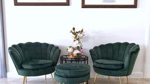 Deux fauteuils verts, recouverts de tissu, avec des pieds dorés, une petite table ronde ornée d’un vase de fleurs et deux tableaux encadrés accrochés au mur.