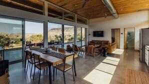 A spacious living area with a large dining table, comfortable seating, and a view of the desert landscape through large windows. - Hawk & Mesa By Homestead Modern (Pioneertown)