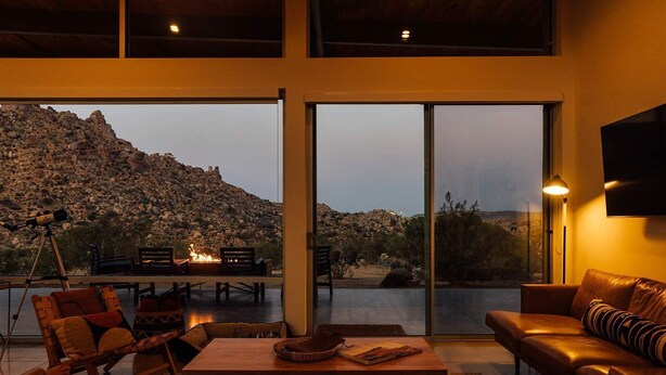 A modern interior with a large sliding glass door leading to an outdoor terrace, a leather sofa, and a dining area with a fire feature. - Hawk & Mesa By Homestead Modern (Pioneertown)