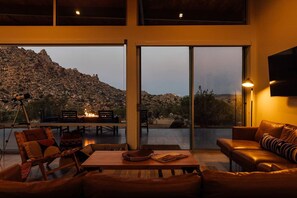 A modern interior with a large sliding glass door leading to an outdoor terrace, a leather sofa, and a dining area with a fire feature. - Hawk & Mesa By Homestead Modern (Pioneertown)